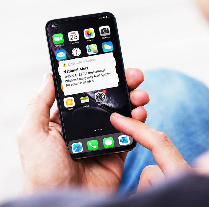 A person holds a mobile device showing a test emergency alert reading, "National Alert. This is a test of the National Wireless Emergency Alert System. No action is needed."
