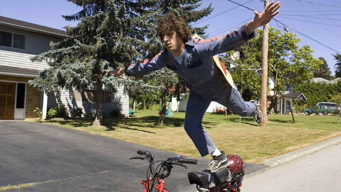 Still from Hot Rod. Wearing a blue jumpsuit, Andy Samberg is standing up with his arms spread out, balancing on top of a dirtbike in the middle of a suburban street.