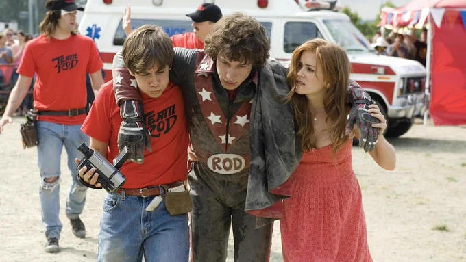 Still from Hot Rod. Jorma Taccone and Isla Fisher are holding up Andy Samberg, wearing a star-spangled jumpsuit and hunched over like he's just been injured.