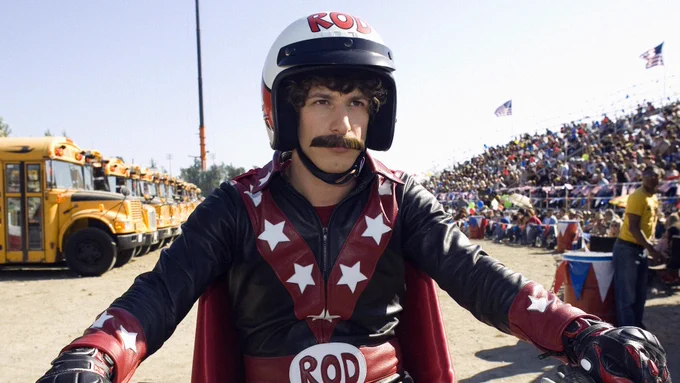 Still from Hot Rod. Andy Samberg is sitting on a dirtbike, wearing a star-spangled jumpsuit that says "ROD" on the belt buckle and on a big motorcycle helmet. He has a fake mustache. Behind him we see a bleacher full of cheering spectators and a row of school buses.