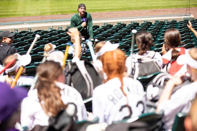 Ryan McMahon sits on the ledge with his back to the field and talks to a group of young campers at softball camp.