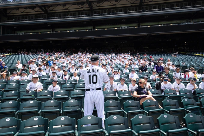 Bud Black stands in his Rockies white pinstripe uniform with his back to the camera while he talks to campers.