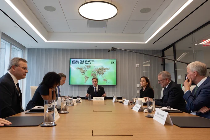 Secretary Blinken sits at a long wooden conference table with stakeholders. A screen behind him shows a world map over a green background and the words "Vision for Adapted Crops and Soils."
