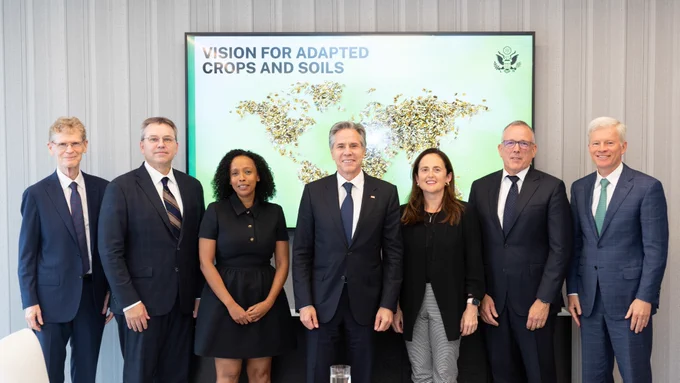 Secretary Blinken stands with a group of agricultural industry and labor leaders and poses for a photo in front of a screen showing a world map over a green background and the words "Vision for Adapted Crops and Soils."