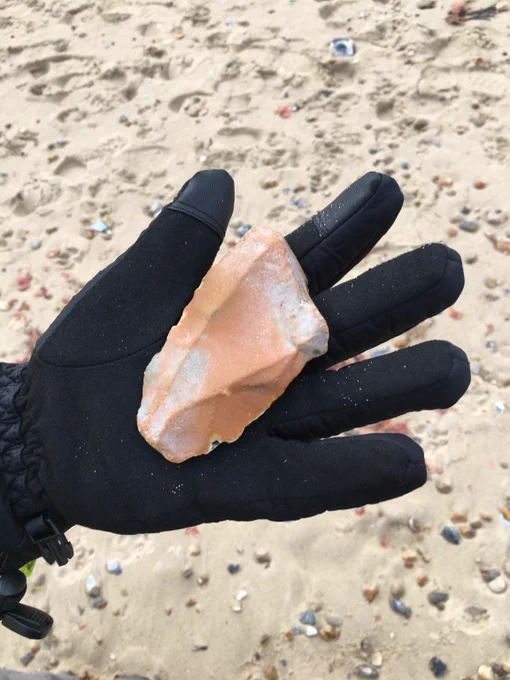 A gloved hand holding a humanly worked piece of flint on a background of beach sand. An example of a Palaeolithic artefact found on the beach after completion of the Sandscaping project at Walcott. This is a Levallois flake (dorsal view) and is stained and rolled, it also shows recent edge damage, most likely as a result of the dredging and beach replenishment process. (Photo: Simon Lewis)