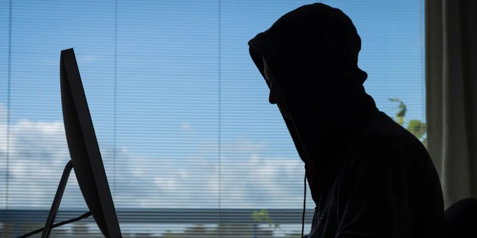 A darkened figure of a person wearing a hood sat in front of a computer with a window with blinds pulled down behind them