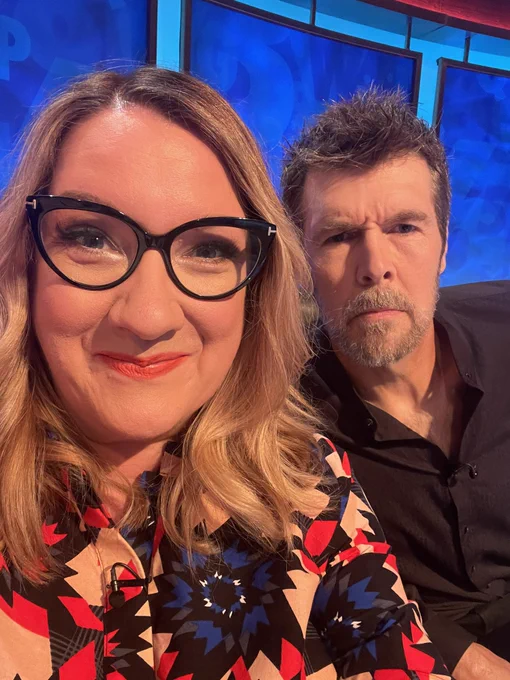 It’s Rhod and I on the set of 8 out of 10 Cats Does Countdown. The background is all blue. I’m on the left wearing a red, blue and grey patterned Zuri dress. My lipstick is red and my hair is blonde and wavy. Rhod is on the right and looking puzzled in a black shirt. We laughed so much.