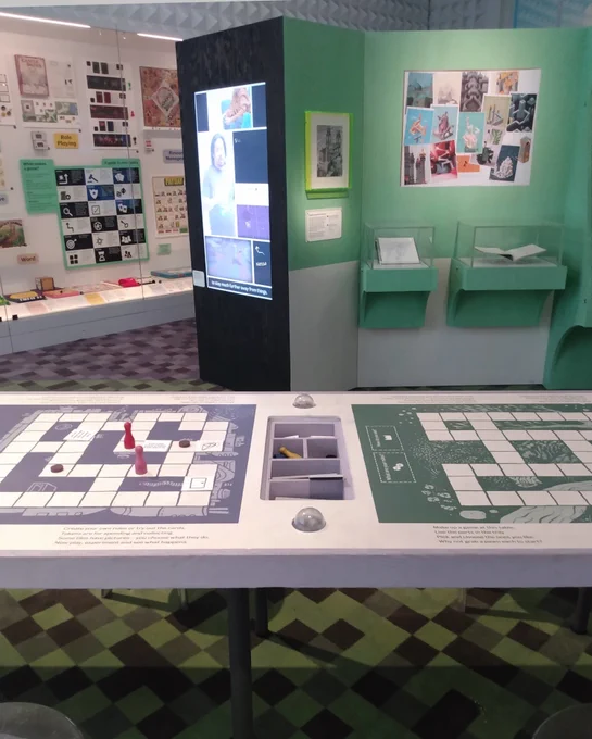 A table printed with two boardgame boards, in an art gallery. A tray in the middle contains assorted game pieces, some of which are in play on the board.