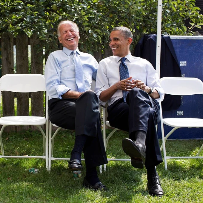 President Joe Biden and President Barack Obama sit beside each other, laughing. 