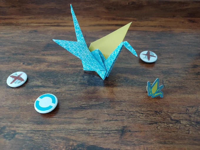 A blue and yellow folded paper crane sits on a shelf with four small buttons -- 2 with a red X over a nuclear weapon, 1 with a Global Zero logo (a blue logo in a zero shape), and one of a folded crane.