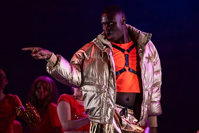 A man wears a gold bomber jacket and shorts and lipsyncs during a scene
