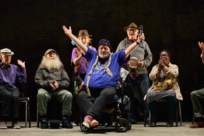 A man in a wheelchair stands centre stage with his arms open. Others sit behind him in a scene that takes place in a care home.