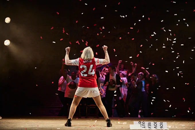 A woman wears white shorts and a red jersey top with number 24 and Green written in white. She cheers as confetti falls and a group of people watch her from the background.