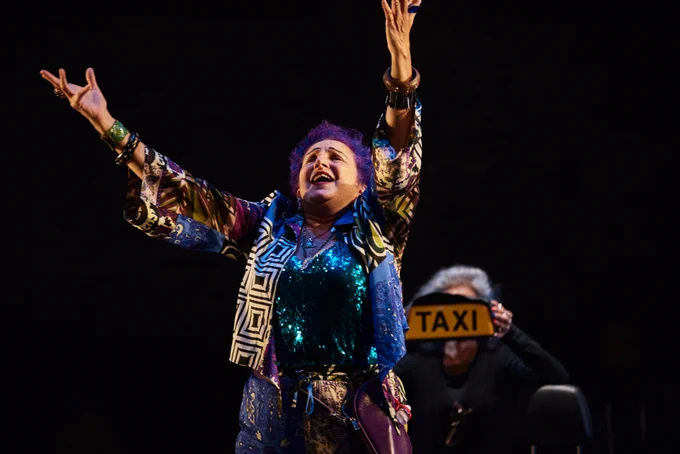 An older woman with purple hair and colourful clothes stands on stage with both arms up.