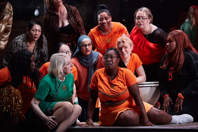 A group of women sit on stage during a scene that takes place in a bathroom.