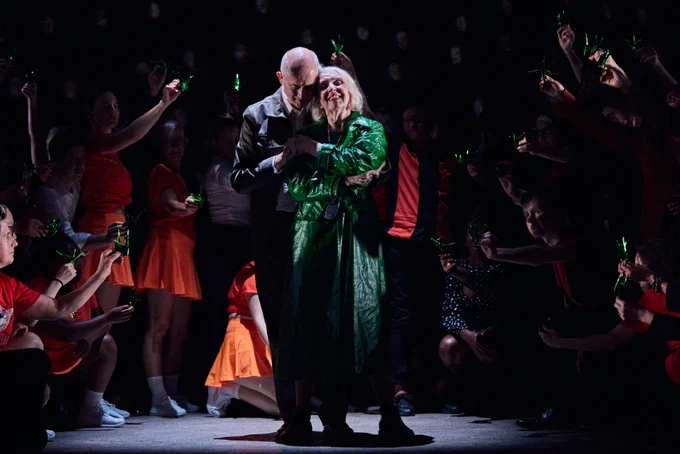An older couple stands together centre stage as other cast members hold green origami birds around them.