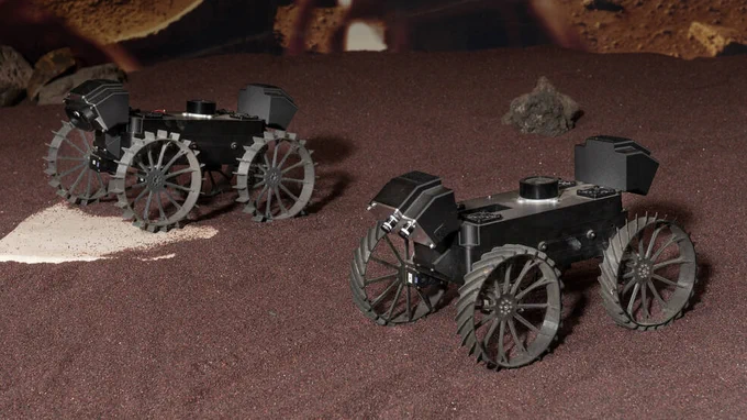 A pair of plastic prototypes of the CADRE rovers demonstrate driving in formation during a test at NASA's Jet Propulsion Laboratory in 2022. The protoypes are black and have four wheels. They sit on simulated reddish Martian soil. Seven of these “Mercury 7” prototypes were built, each named for one of NASA’s seven Project Mercury astronauts. John (for John Glenn) and Scott (for Scott Carpenter) are shown here. Credit: NASA/JPL-Caltech