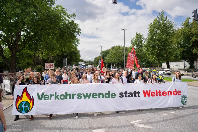 Großes Banner auf dem Verkehrswende statt Weltende mit einer brennenden Erde steht. Dahinter Menschen mit Schildern, Fahnen und Bannern auf einer Straße. Rechts und links sind Bäume und teilweise Autos zu sehen 