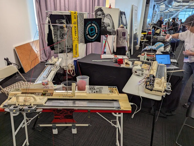 Four tables featuring different electronic knitting machines from the 1980s. In the background hung on easels are a few samples including Neil Armstrong on the moon, a scarf, and the "We can do it!" wartime poster.