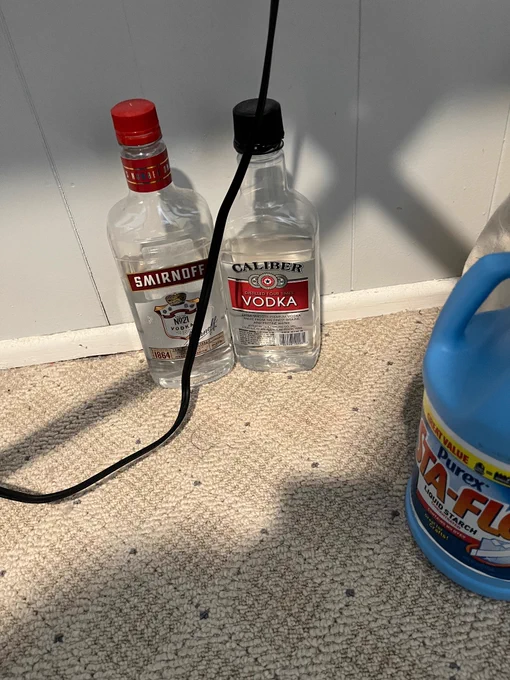 Two bottles of cheap vodka on the carpeted floor underneath the ironing board