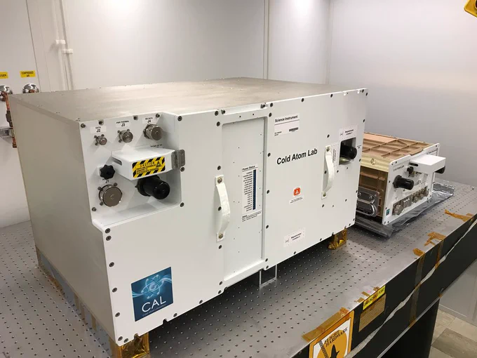 A rectangular white metal box is shown on top of a steel table. The box contains the label "Cold Atom Lab" to the far right of it, next to one of two white handles. To the left of the box is an indentation with room for three knobs of small to larger sizes, a rectangular vent with a "caution" label on top and then two larger knobs in silver and black right underneath. At the very bottom on the left side of the metal box is a sticker with an atom illustration in various shades of blue and the words "CAL Cold Atom Lab" in white.