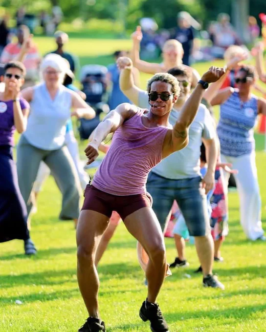Photo of a dancer in a purple tank top and shorts and sunglasses in a leaned pose with one arm up. Behind them is a crowd of people following along.
