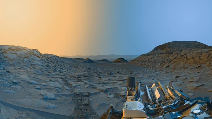 Two panoramas taken in the morning and afternoon on Mars are combined to create a color-enhanced landscape image. Both panoramas of the area, known as "Marker Band Valley," were taken by the Curiosity Mars rover's navigation cameras. The detailed photo shows yellow coloring in the sky to mark the afternoon hours and it transitions to a blue color to indicate the morning color. The taupe-colored landscape shows a large, rocky hill to the right and smaller rocky hills in the distance toward the center and out into the horizon. To the left is a rocky outcrop and as we get closer to the foreground the area shows rover tracks along the sandy floor. To the bottom-right corner, the backend of the off-white, dusty rover is shown. Part of the rover's wheel can be seen as well as three of its antennas. The Radiation Assessment Detector, or RAD instrument, appears as a white circle in the lower right portion on the back of the rover.