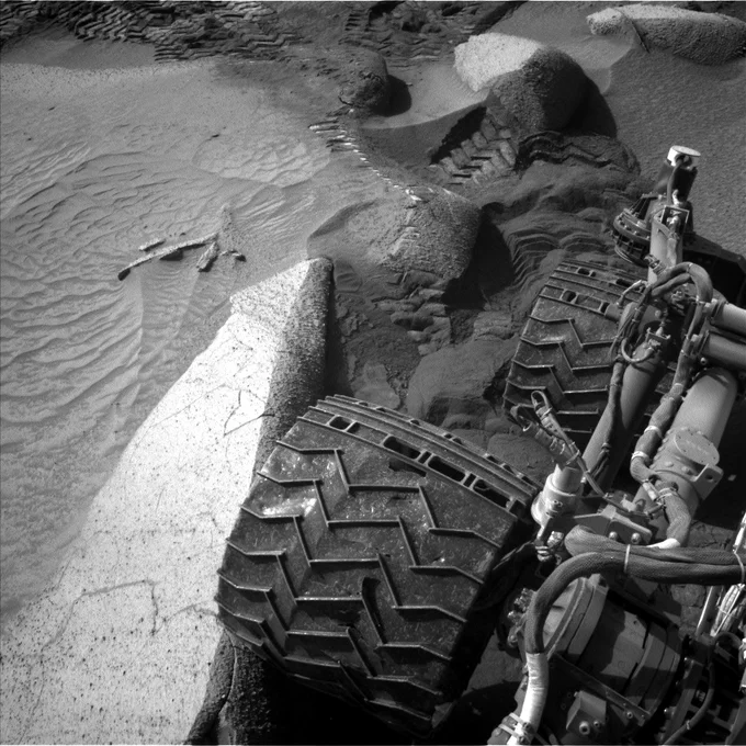 A black-and-white square image shows the wheels of the Curiosity rover as it navigates a tricky trail on Mars. The wheels of the rover take up a big portion of the image toward the right and part of the wheel connections are shown with wires and zip-ties. To the right, the Martian surface is very sandy with ripples and large rocks. One of Curiosity's wheels is against a big rock and behind the rover are tracks deep in the sand, showing the slippery terrain it had to deal with.