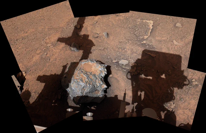 This composite image shows Curiosity's view of its Martian workspace. A large rock is centered between shadows the rover cast onto the surface. Curiosity's "head" is to the right of the rock, and its arm is to the left. The rock itself is roughly a foot in diameter and mostly round, with divots across its rough surface. It is gray/silver. Mars dirt has settled on portions of it. In the rest of the image, the light red Martian surface is sprinkled with smaller rocks and pebbles.