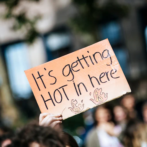 Cardboard sign raised above an out-of-focus crowd. Sign reads "It's getting hot in here."