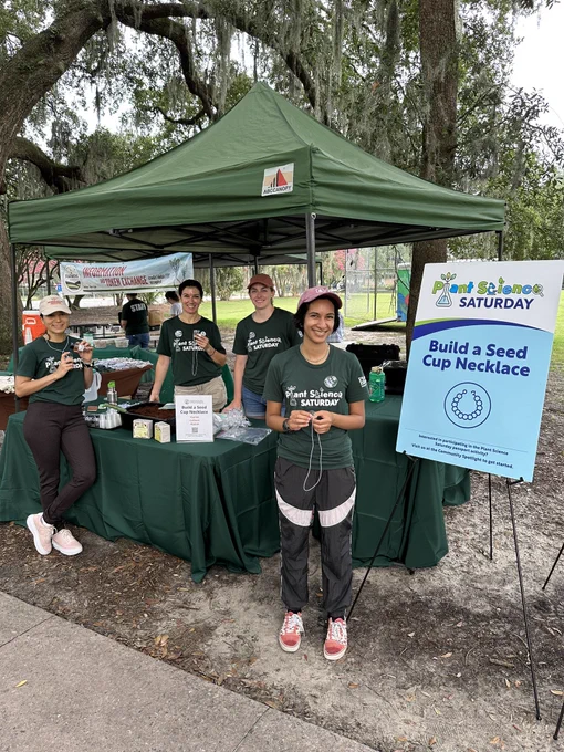 Four smiling volunteers posed for a photo next to the Build a Seed Cup Necklace sign!