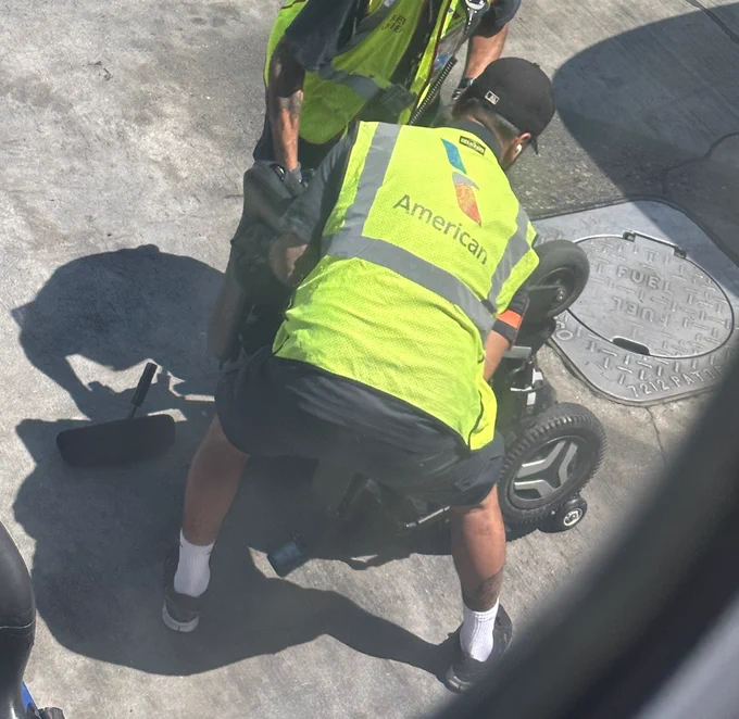 Wheelchair flipped over with pieces strewn on the ramp at an airport, American Airlines staff crowded around it.