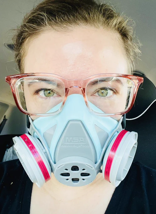 A white woman with brown hair pulled back in a bun wearing a pastel blue and gray elastomeric respirator with hot pink bands on the filters, and coral clear plastic stoggles.