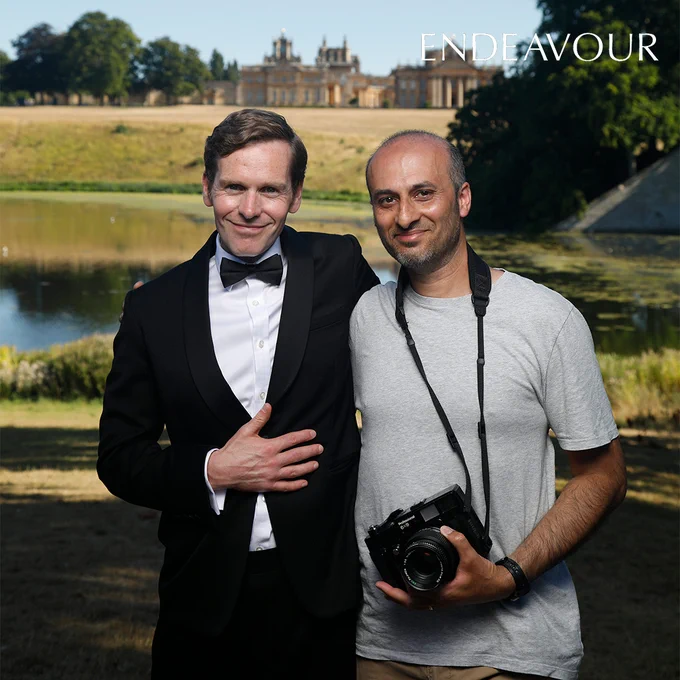 Actor Shaun Evans, wearing a tuxedo, is stood alongside a member of the production team, who is dressed casually and holding a professional digital SLR camera. In the backdrop is a grand stately home.