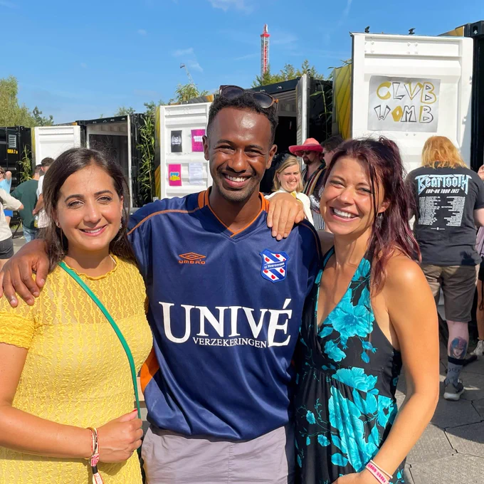 Foto met links Songul, die een kanariegele jurk draagt. Habtamu in het midden met een blauw SC Heerenveen-voetbalshirt en ik sta rechts voor de kijker met een zwarte jurk met lichtblauwe bloemen. Op de achtergrond zie je een paar mensen die met de rug naar ons toe staan. Foto is buiten, voor een aantal containers waarin de experimenten plaatsvinden.