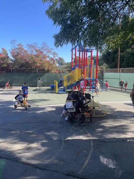 Playground with a jungle gym, child riding a tricycle and a child in a motorized wheelchair. Many children playing.