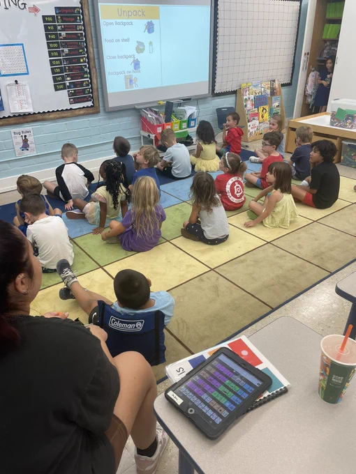 Four year old children in a transitional kindergarten class sitting on the carpet for their morning meeting. An adult classroom aide sitting next to an iPad displaying communication software for use by a student who is non-verbal.