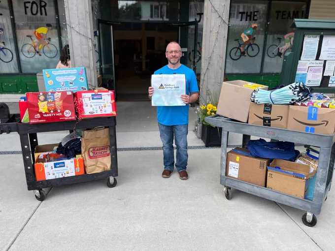 Man in blue shirt holding a Middlesex Savings Bank sign while standing between two carts full of pantry donations outside of our Maynard location.