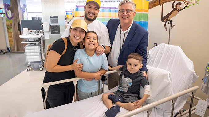 Four adults stand around a hospital bed where a young boy is connected to an intravenous line delivering life-altering therapy