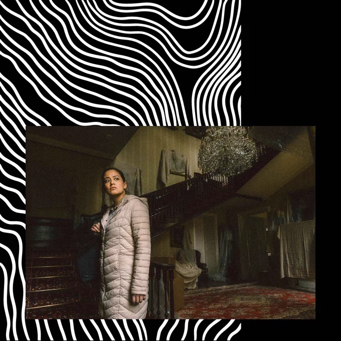 [Image: A woman standing in an old mansion foyer, looking into the distance]