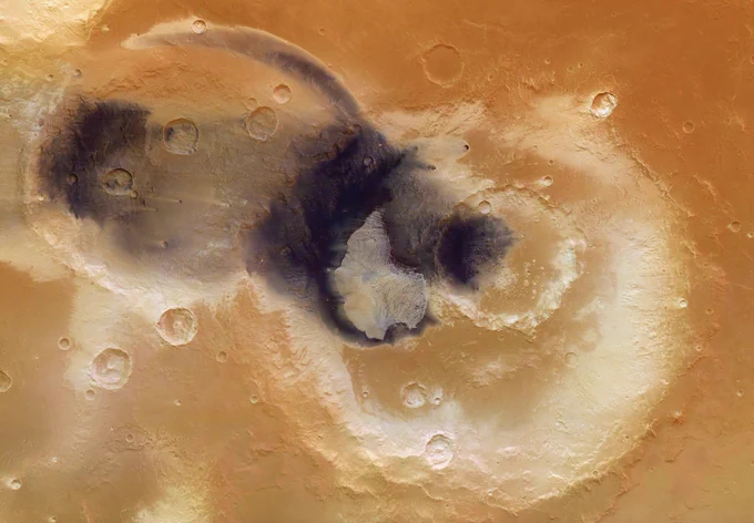 A mound of light-coloured sulphate deposits formed from evaporating water sits inside the Becquerel crater on Mars amid a sea of dark wind-blown deposits. The darker material has blown towards the south-southwest (top left) of the image in a wide swath and across tiny craters there – their raised rims protect the material immediately downwind from being swept away.