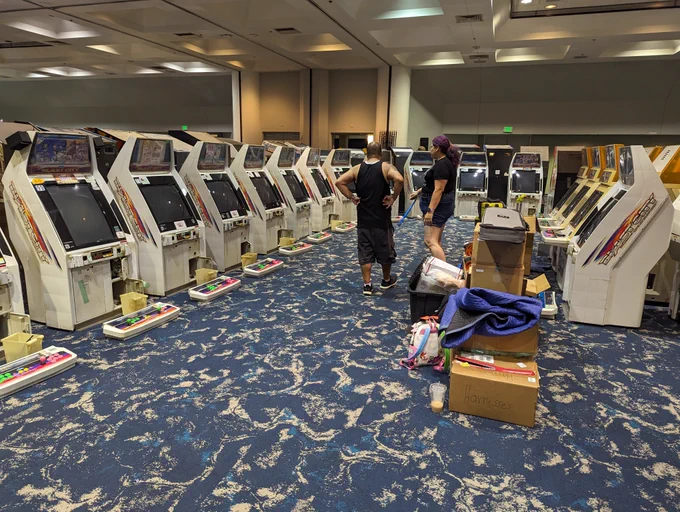 
A row of at least 14 Japanese Sega Blast City cabinets wrapping around the frame of the photo with two people setting up in the middle. Most of the control panels are lying on the floor ready to be attached.