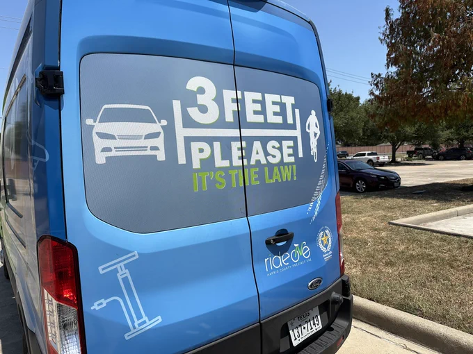 Picture of a van with the message on the back that says “3 feet please. It’s the law!” Showing an image of a car and a bike spaced apart.