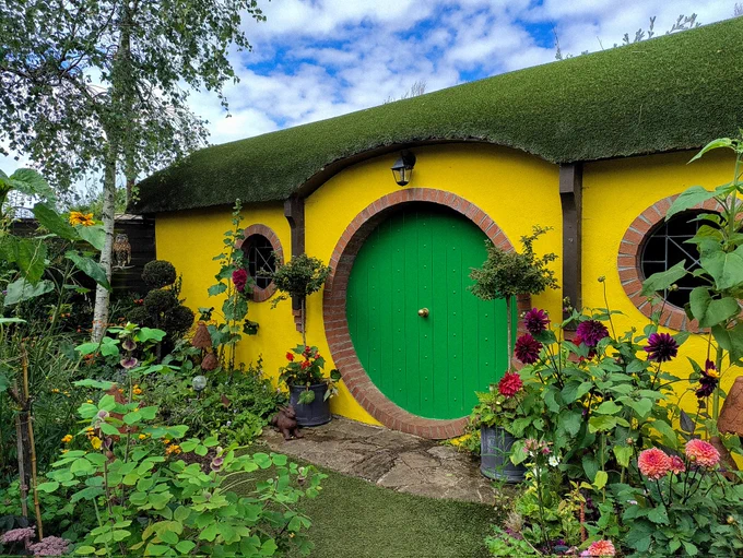 photo of a hobbit style bright yellow cottage with a round green front door and a green moss roof surrounded by plants and flowers 