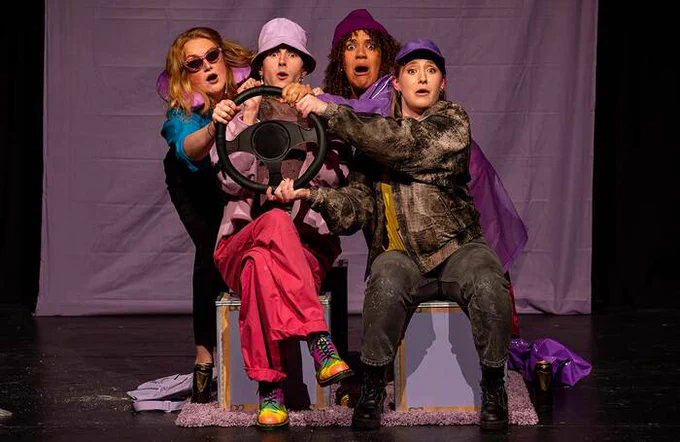 Four performers on stage behind a steering wheel with shocked facial expressions