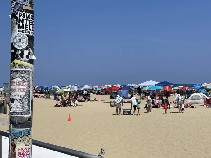 Beach scene. Lots of people and umbrellas. A pile to the left is covered with pasted-on ADs for local events.
