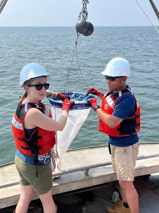 This photo shows SERC researchers Kristina and Clinton on a boat conducting Zooplankton surveys in Chesapeake Bay. They're both holding onto a plankton tow net and wearing white hard hats and red PFDs. PC: Brenda María Soler Figueroa