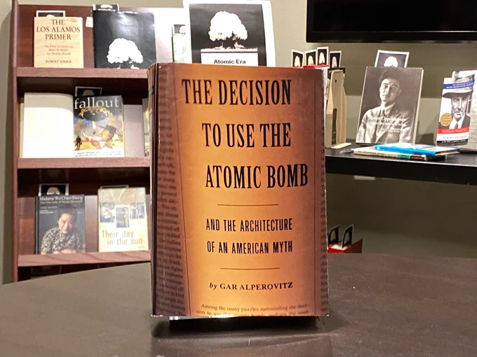Photo of book on table with shelves of books and book cart in background (all on atomic era topics). Book featured is "The Decision to Use the Atomic Bomb."