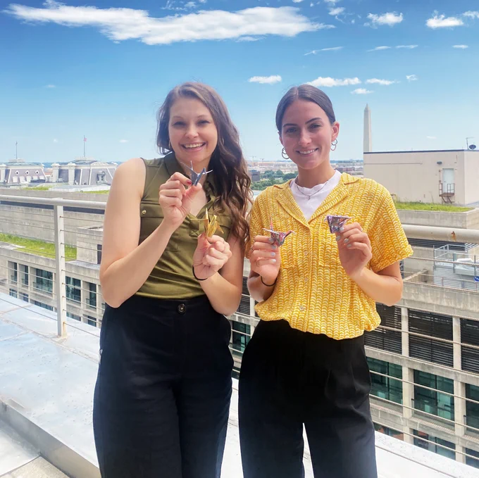 Two women hold colorful origami cranes against the backdrop of the skyline in Washington, D.C.