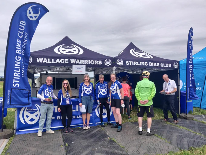 A blue vertical banner on the left, two blue gazebo tents in the centre, with five people in blue Stirling Bike Club jerseys looking at the camera. One visitor in a green jacket talks to a man in a white shirt.